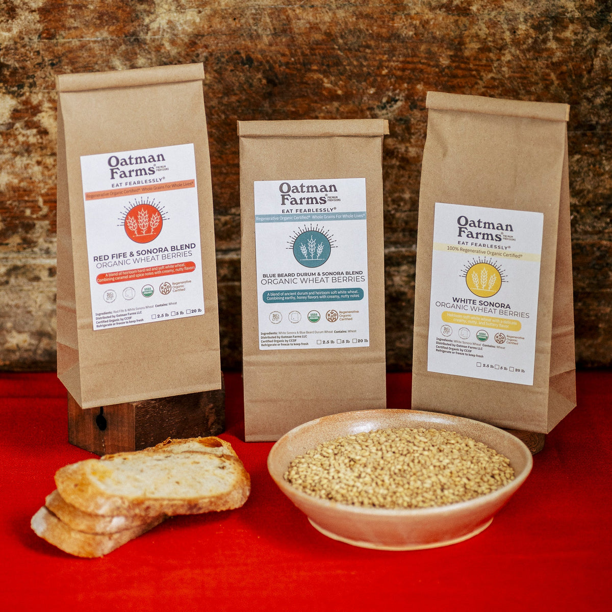 photography of three closed bags of Oatman Farms wheat berries on a red tablecloth and with a stack of bread and wheat berries in the foreground.