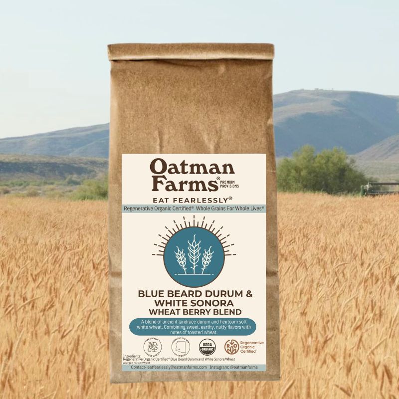 composite image of a brown bag labeled with Oatman Farms heritage wheat berries imposed over a wheat field with mountains in the background.